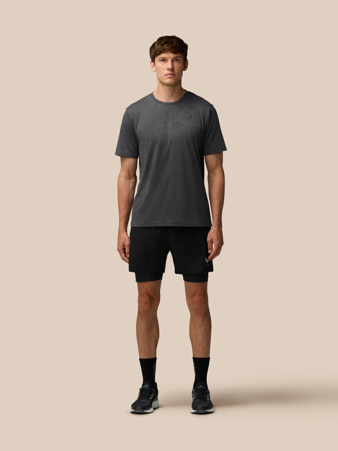 Full body shot of a male model wearing Flow 2-in-1 shorts in black with a gray patterned T shirt against a beige background