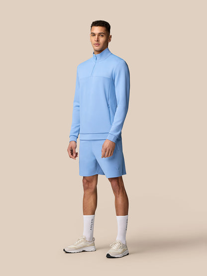Full-body shot of man in sky blue quarter-zip top and matching shorts with white sneakers and socks
