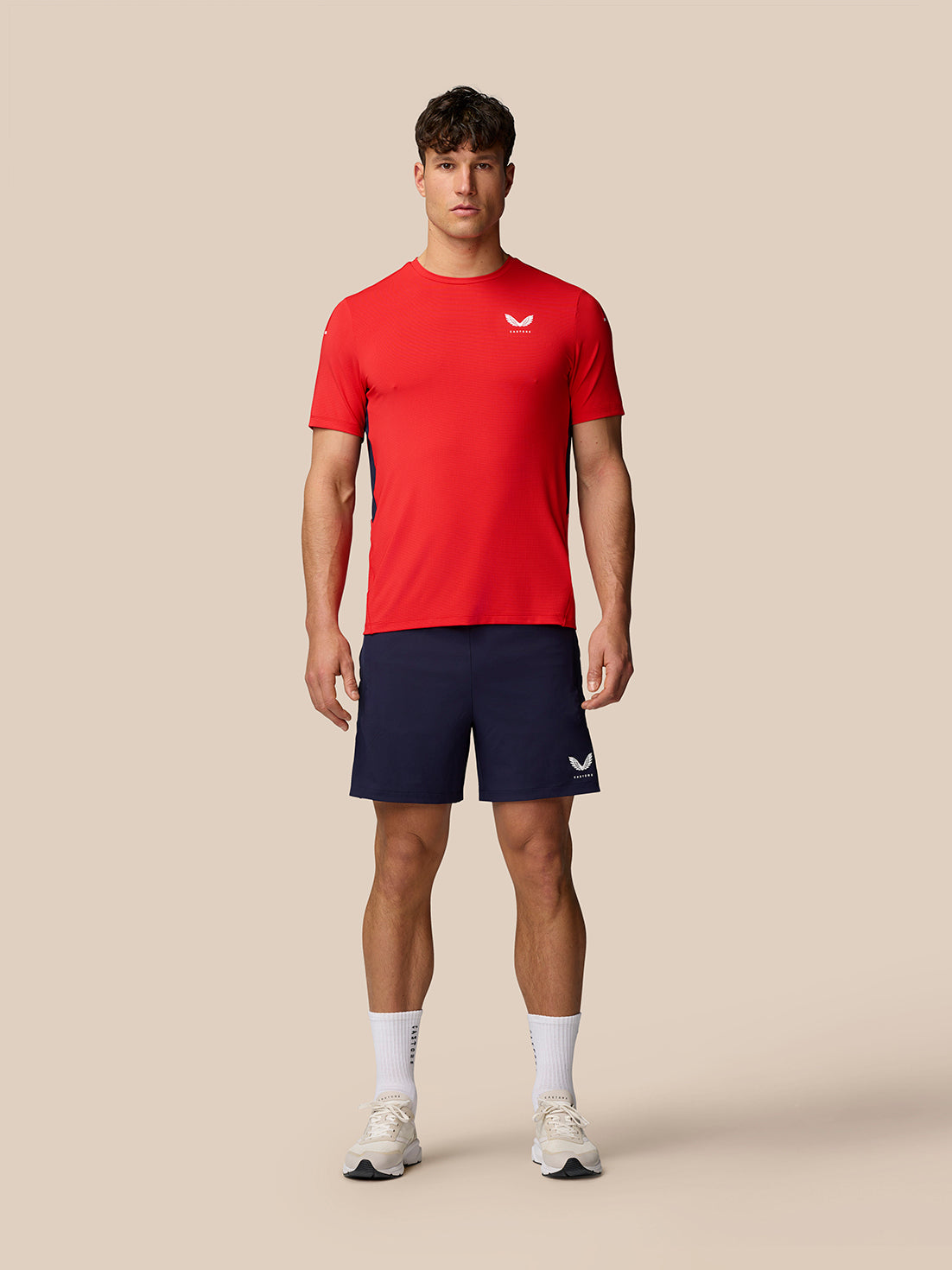 Full-body shot of a male model in a red Apex Training T-Shirt and navy shorts, featuring white Castore branding on the chest.
