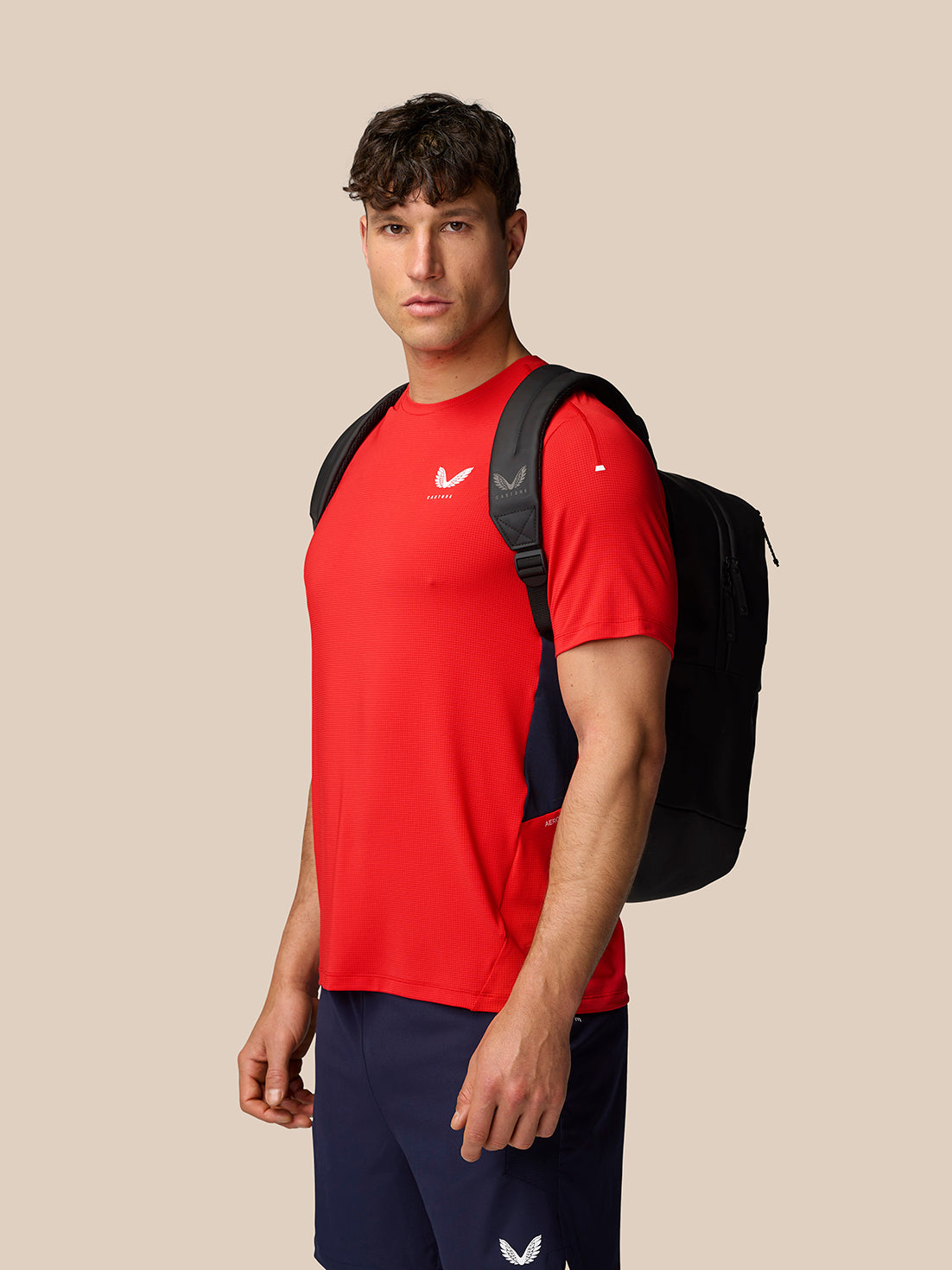 Male model wearing red Apex Training T-Shirt with navy shorts and backpack, side profile.
