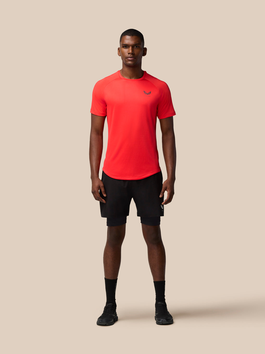 Male model wearing Flow Training Muscle Grip T Shirt in bright red with black shorts against a neutral beige backdrop