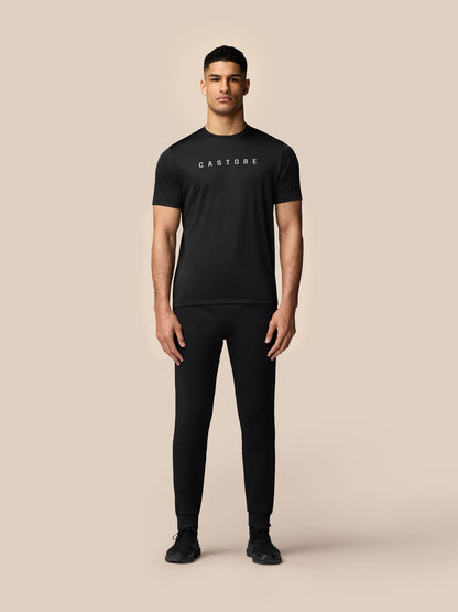 Full body view of a man wearing a black graphic training T shirt by Castore with black joggers against a beige background