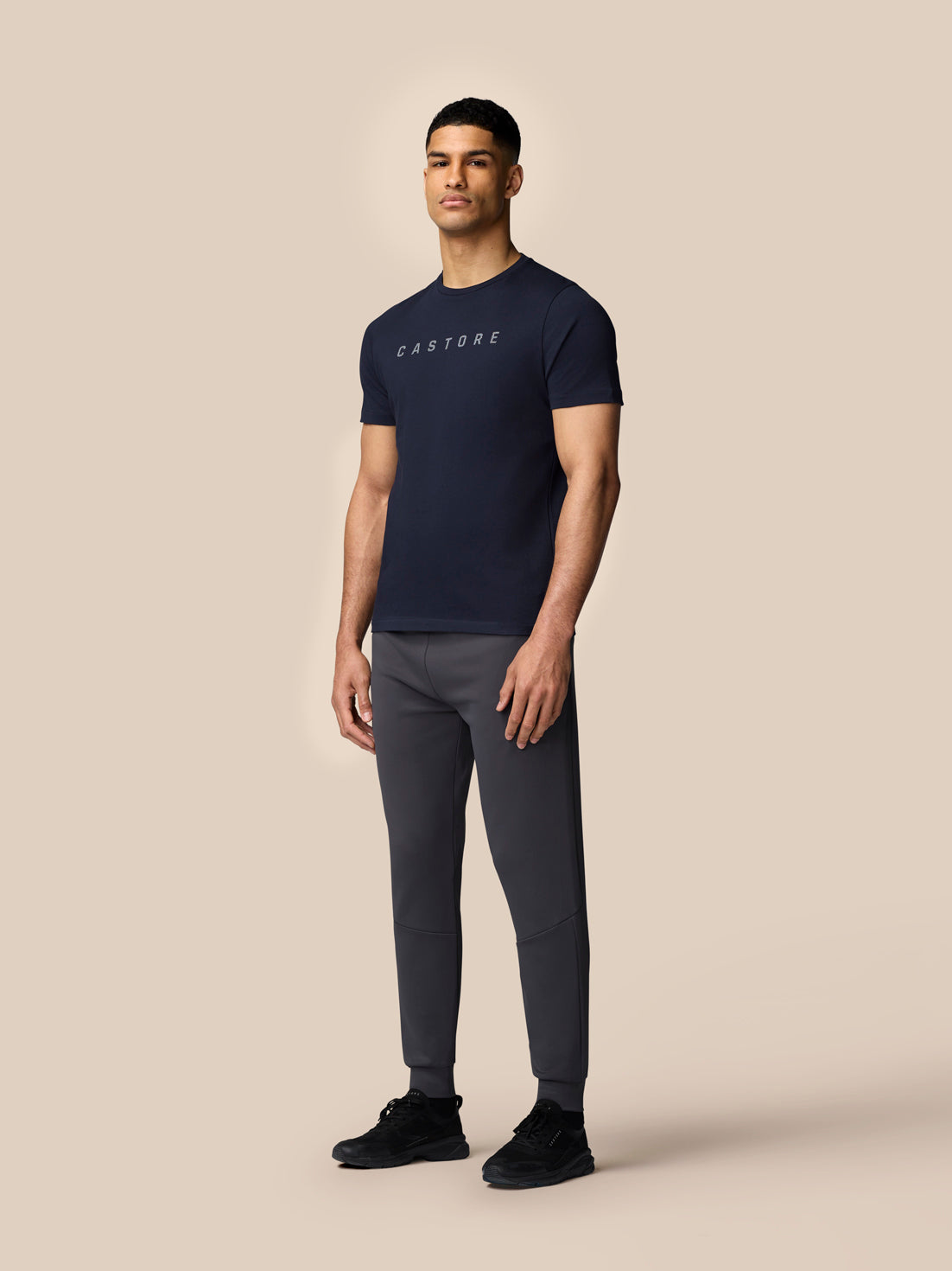 Full-body view of a man in a navy graphic Castore T-shirt with drop shoulders, paired with gray joggers and black sneakers.