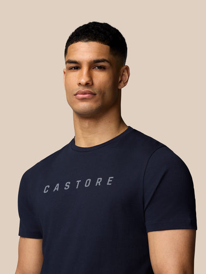 Close-up of a man in a navy Castore graphic T-shirt with crew neck and chest logo against a beige background.