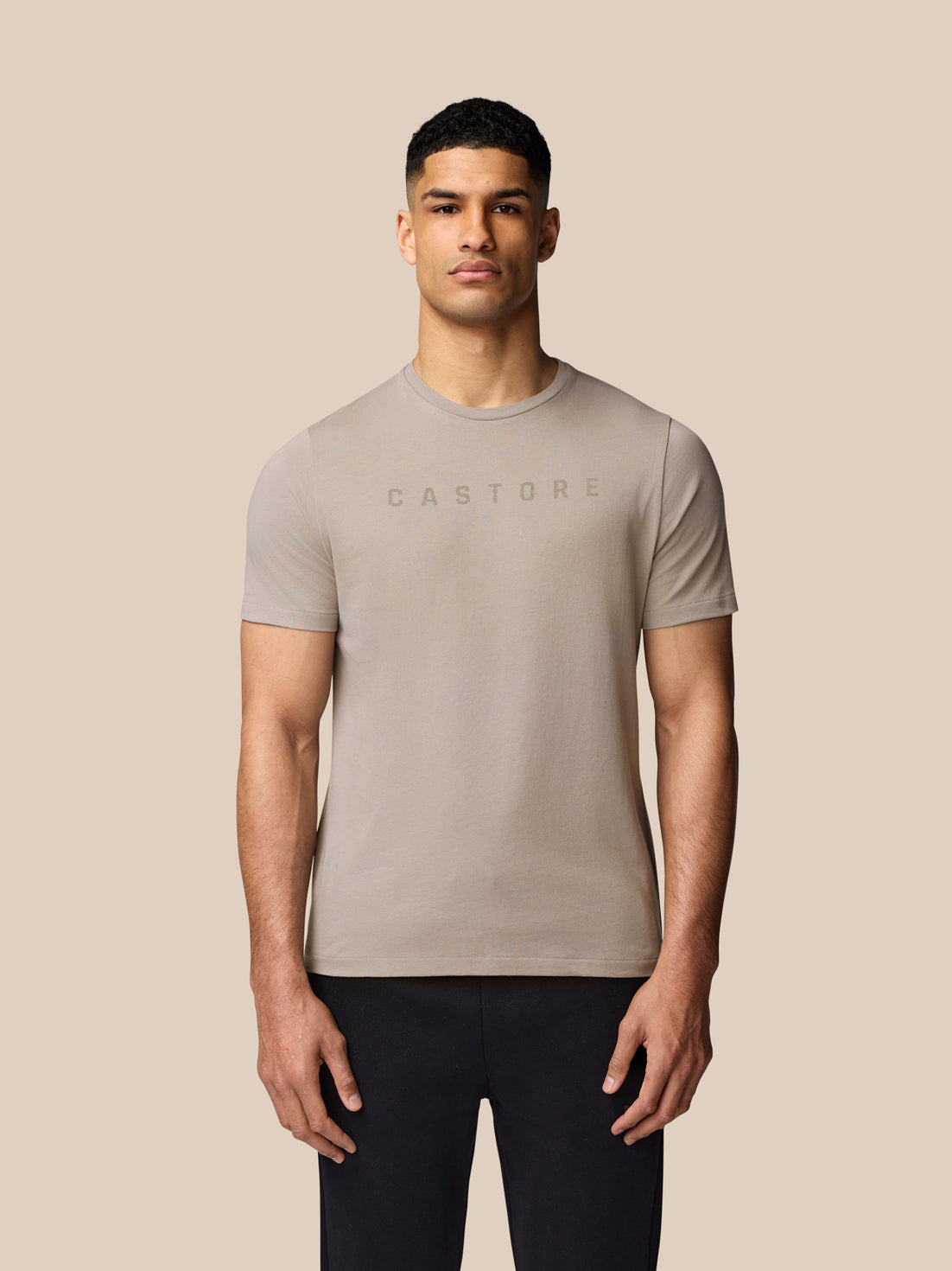 Portrait of a male model wearing the Rock Grey graphic T shirt with Castore logo on a beige backdrop.