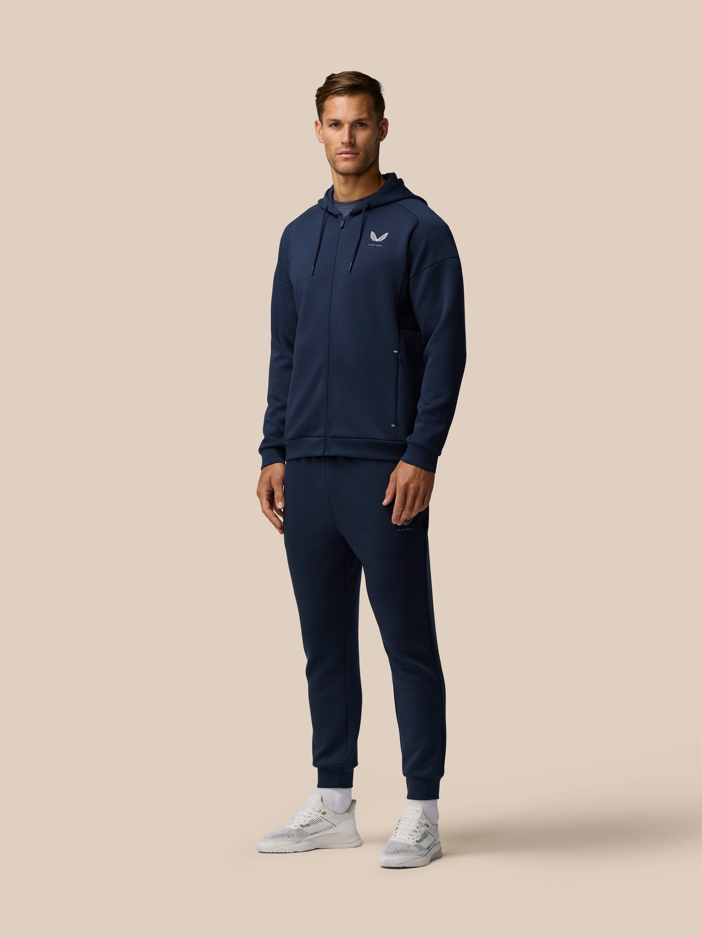 Front view of a man in navy Travel Lifestyle Zip Hoodie with logo on the chest, wearing matching joggers and white sneakers.