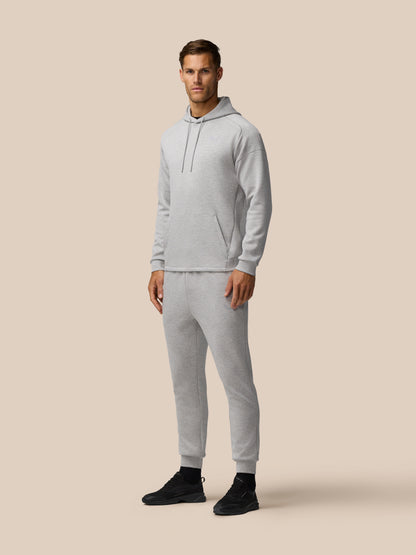 Model wearing light grey marl travel hoodie with hood and front kangaroo pocket against a beige backdrop