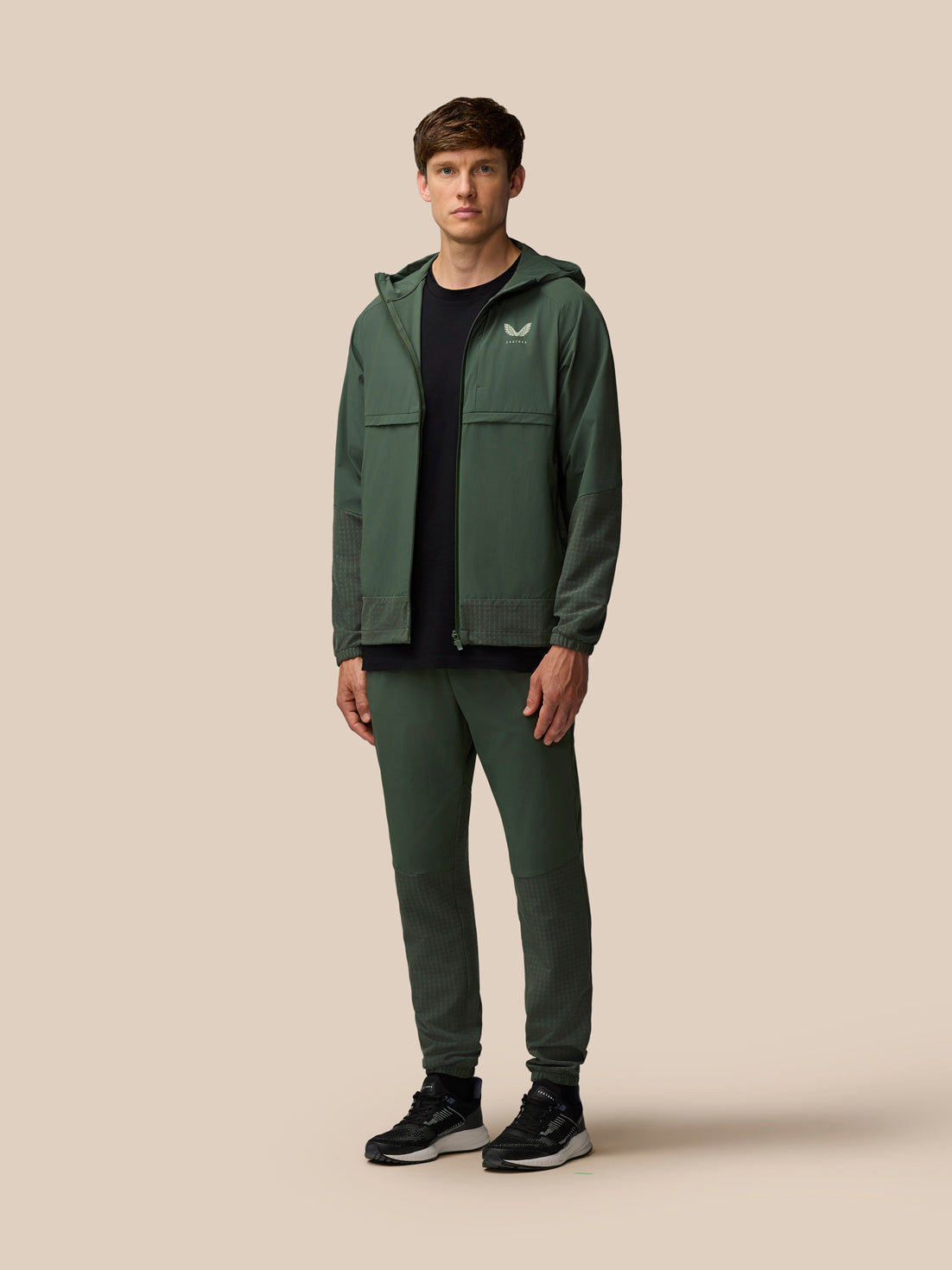 Full-length model in a dark green Castore Flex Winterised Woven outfit wearing a jacket and matching joggers.
