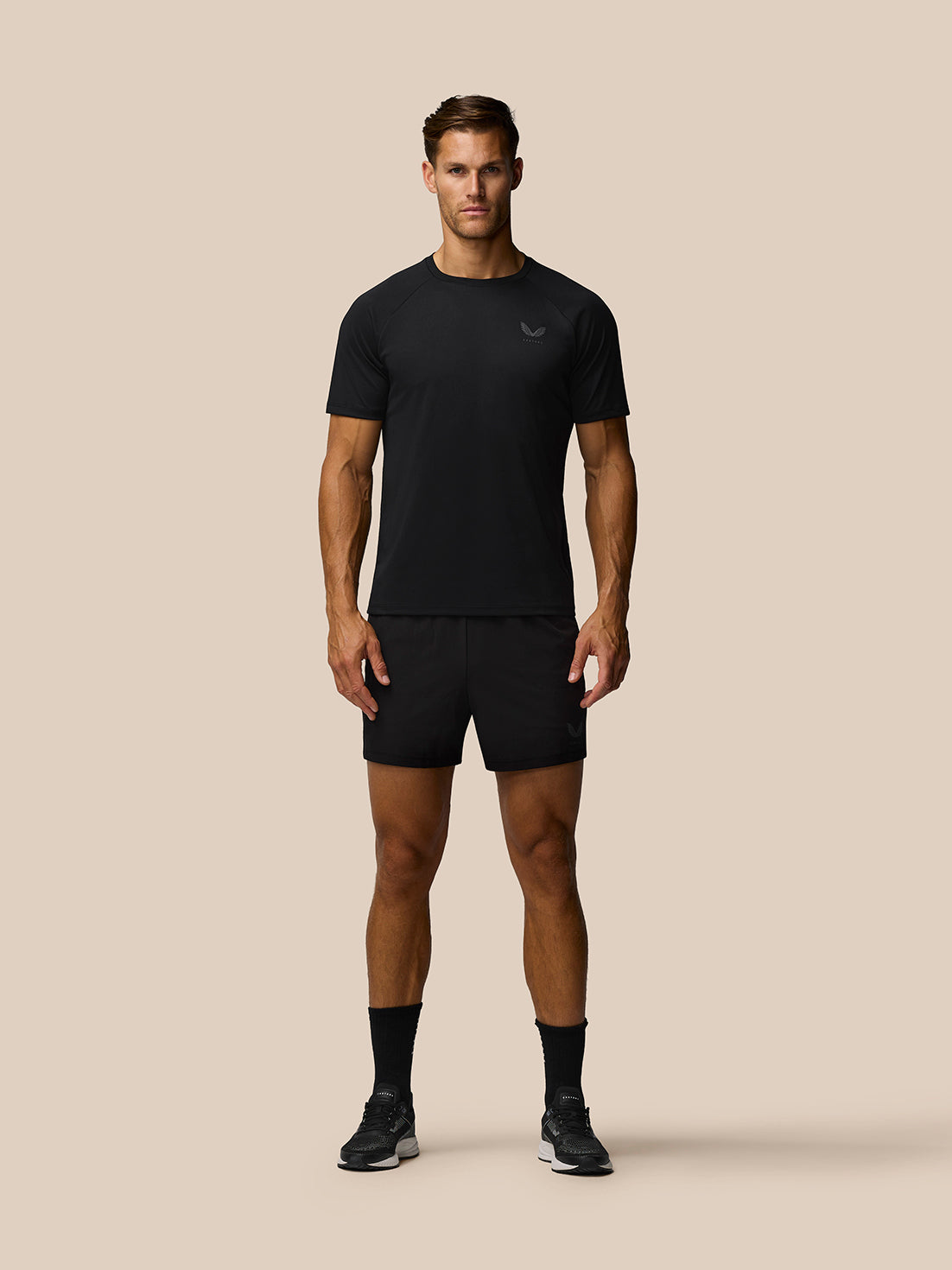 Full-body view of a male model wearing black Adapt 5 Inch shorts with a Castore logo on the leg