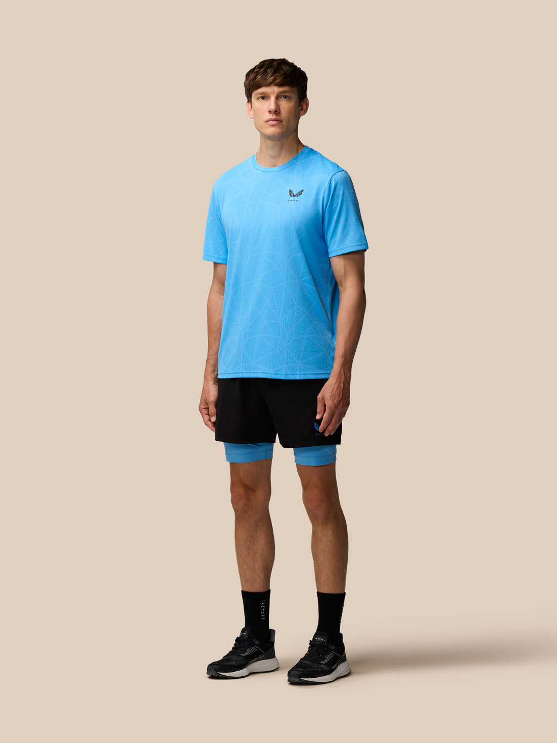Full body view of male model in the blue Flow Training Engineered Knit T Shirt with black shorts and blue leg wraps against a beige backdrop