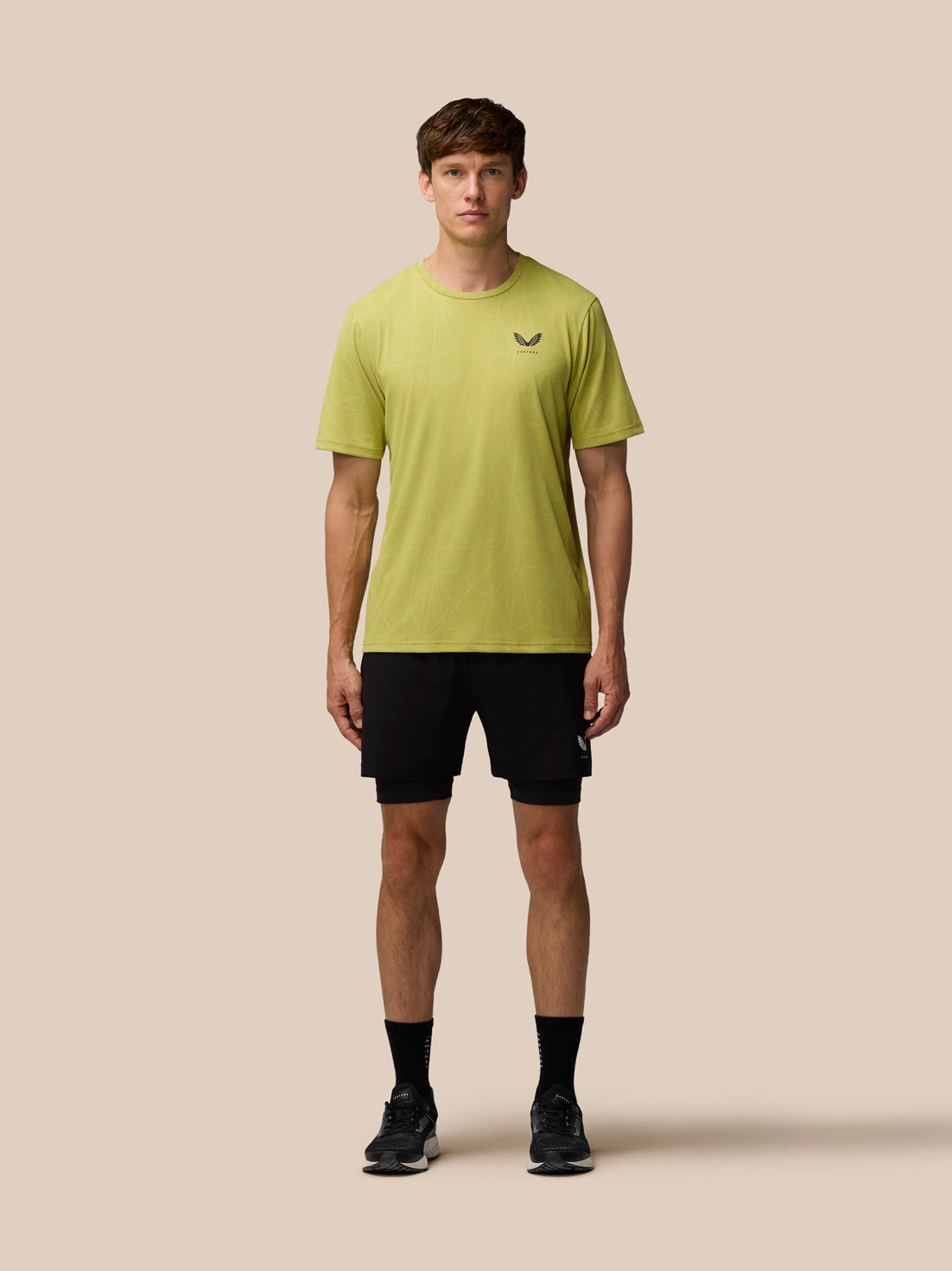 Model wearing Flow Training Engineered Knit T-Shirt in Mustard with black shorts, Castore logo on left chest against a beige background.