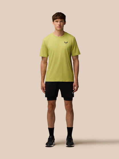Model wearing Flow Training Engineered Knit T-Shirt in Mustard with black shorts, Castore logo on left chest against a beige background.