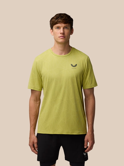 Male model wearing Flow Training Engineered Knit T-Shirt in Mustard with black shorts, Castore logo on left chest, beige studio backdrop.