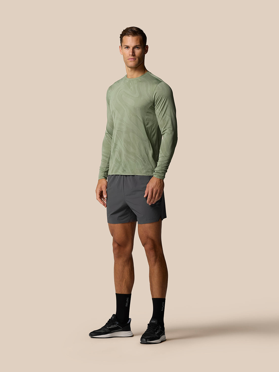 Full body front view of a man in Zone Training LVS Long Sleeve Top Thyme with dark athletic shorts.