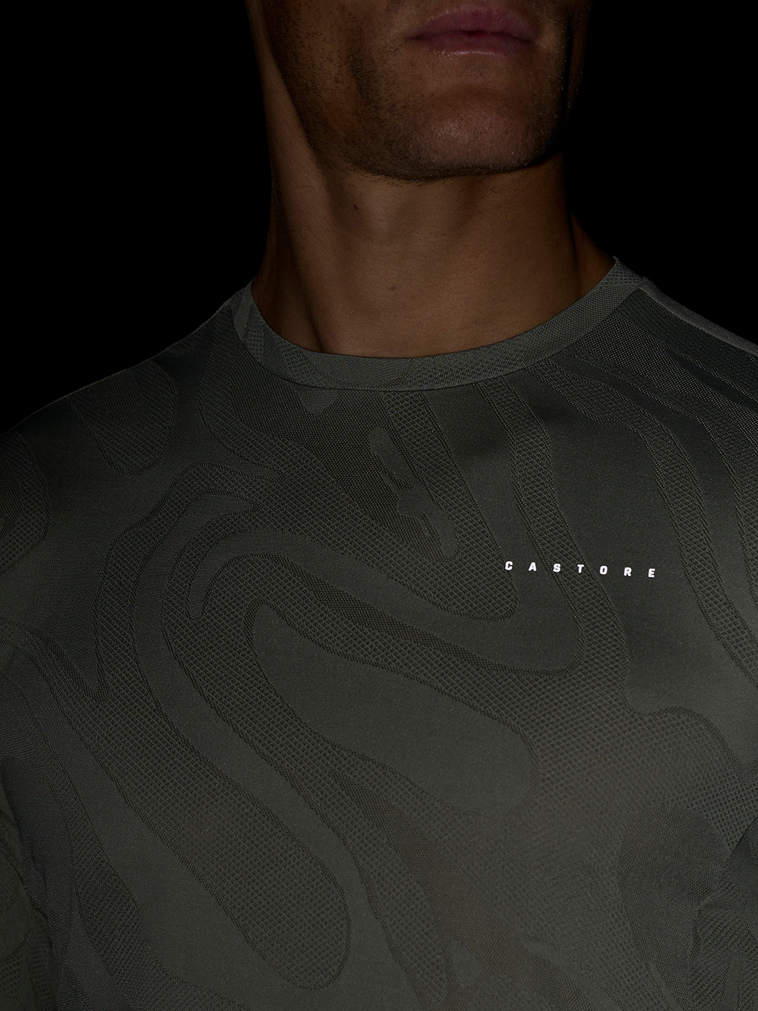 Close-up of a man wearing a dark patterned Zone Training LVS Long Sleeve Top in Thyme with a round neckline and a Castore logo on the chest