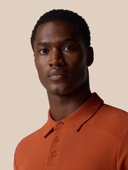 Male model wearing a dark rust polo shirt with a classic collar and three-button placket against a beige backdrop, subtle Castore branding on the chest.