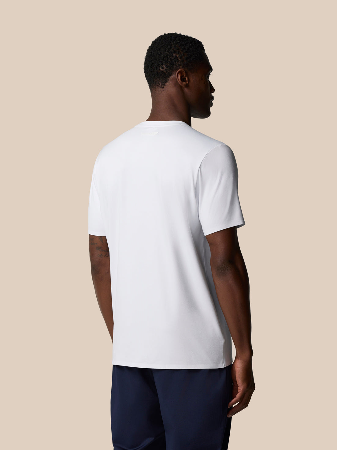 Back view of male model in a white Elevated Lifestyle T‑Shirt against a beige backdrop.