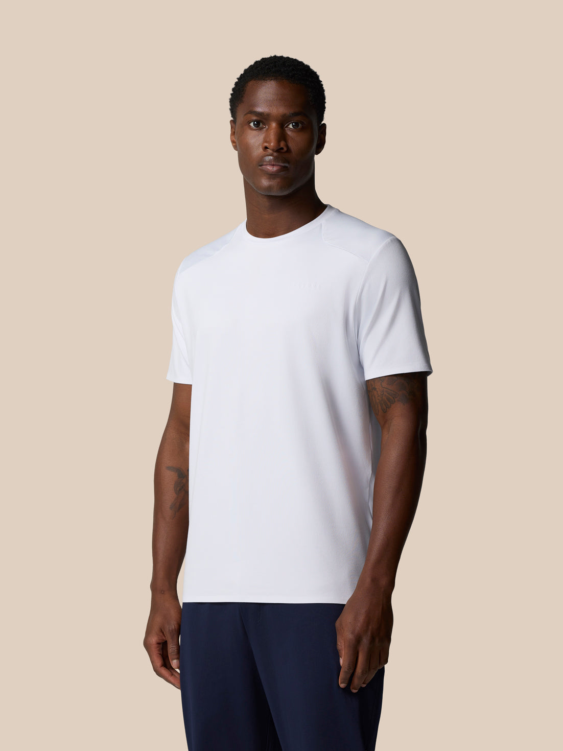Male model wearing white Elevated Lifestyle T‑Shirt with navy shorts against a beige backdrop.