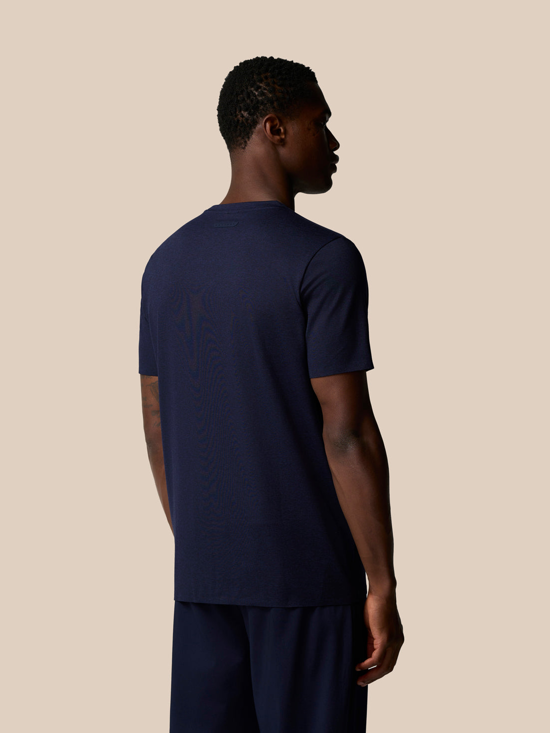 Back view of man in navy Elevated Lifestyle T Shirt against beige background.