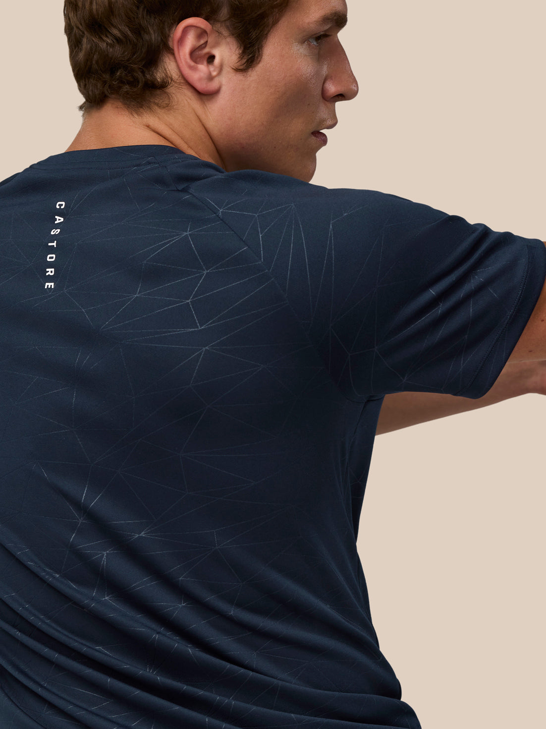 Back view of male wearing navy Adapt All Over Print T Shirt showing vertical Castore wordmark on upper back and geometric print
