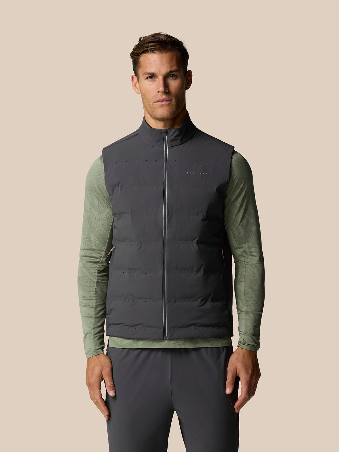 Male model wearing Zone Training LVS Insulated Gilet in gunmetal over olive long sleeve, with front zipper and sleeveless design.