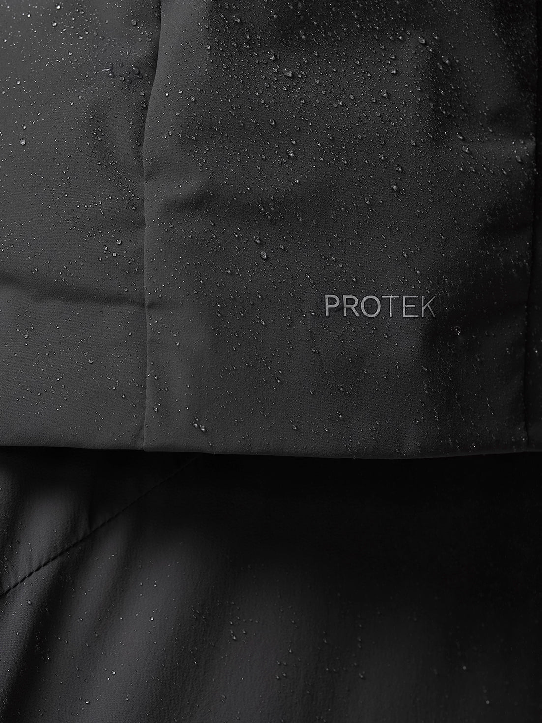 Close-up of dark jacket fabric with PROTEK branding and water droplets on the surface