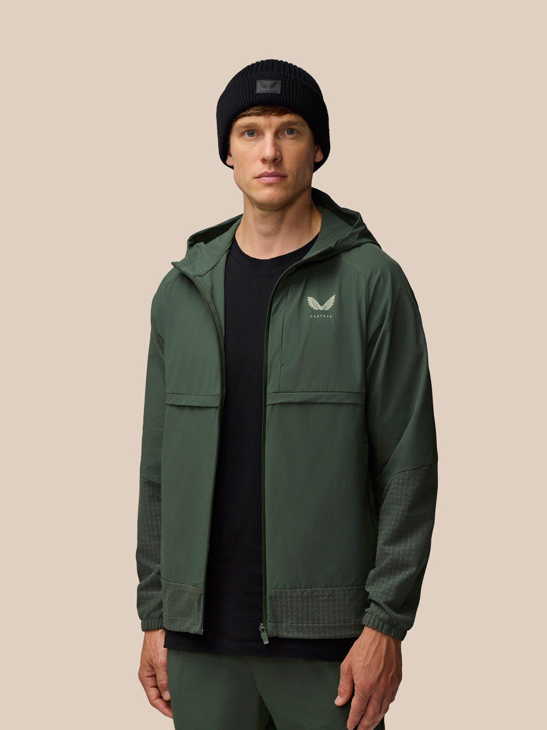 Male model in dark green Flex Winterised Woven Jacket with hood down, Castore logo on left chest, wearing a black beanie.