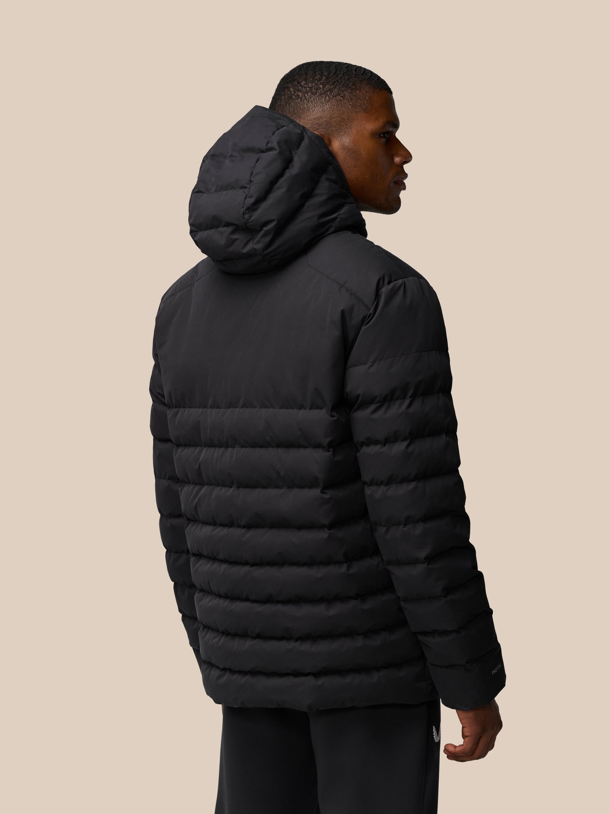 Side profile of a man wearing a black quilted jacket with a hood against a beige backdrop