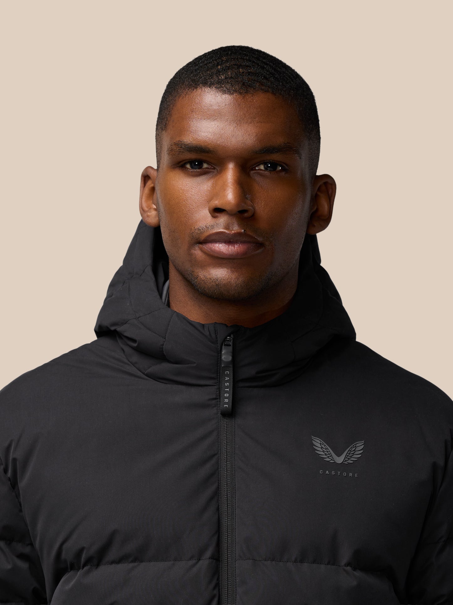 Close-up of a man wearing a black lightweight padded jacket with a hood and front zipper against a beige background.