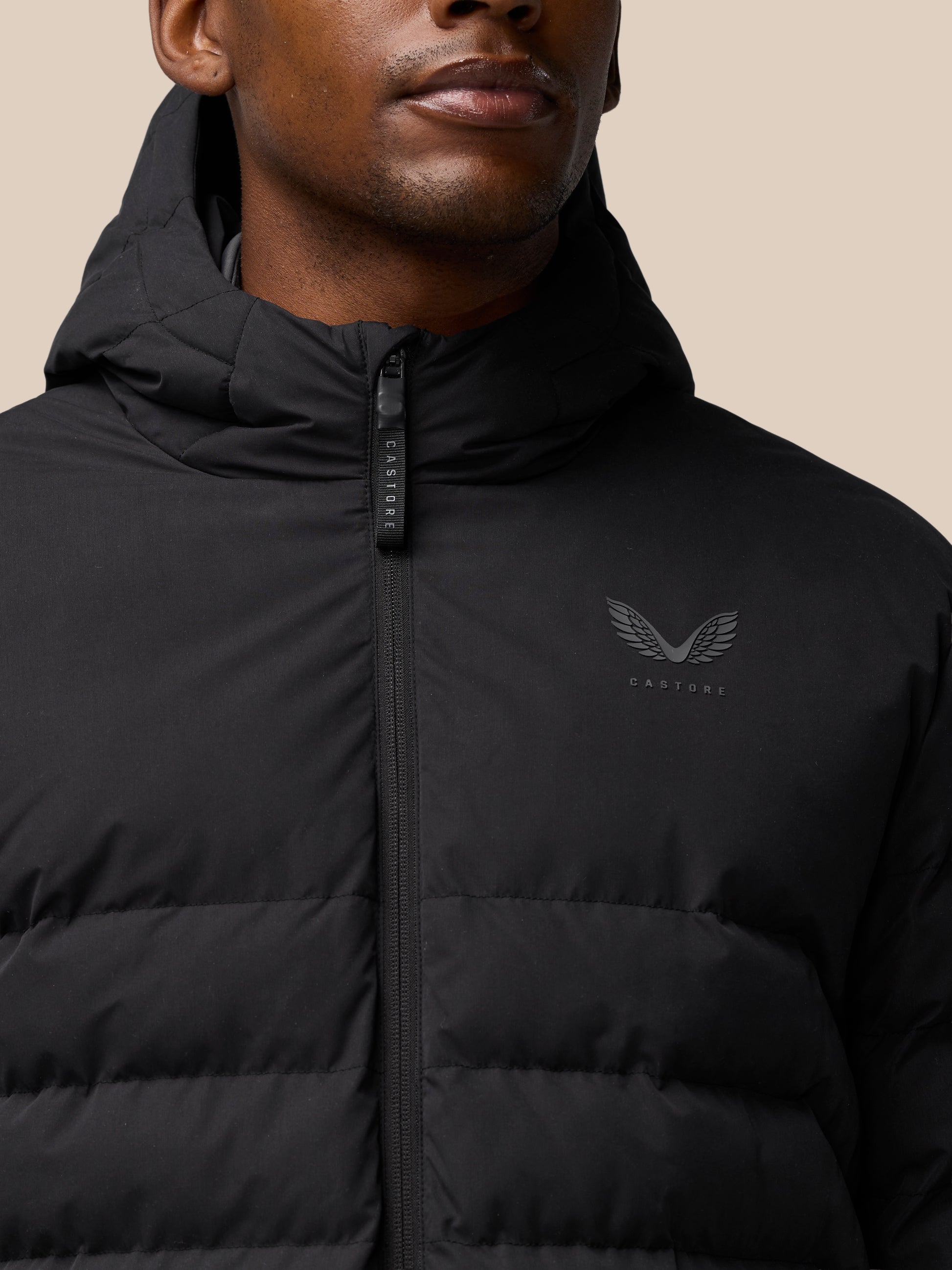 Close-up of a black lightweight puffer jacket with a front zipper, quilted panels, and the winged Castore logo on the chest under a hood