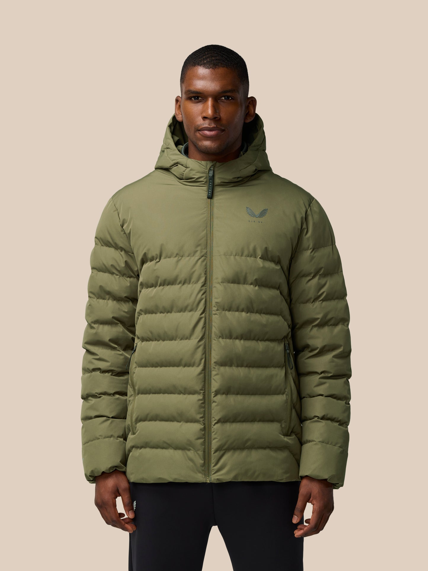 Man wearing ivy green lightweight puffer jacket with hood and front zipper, logo on chest, against a beige backdrop.