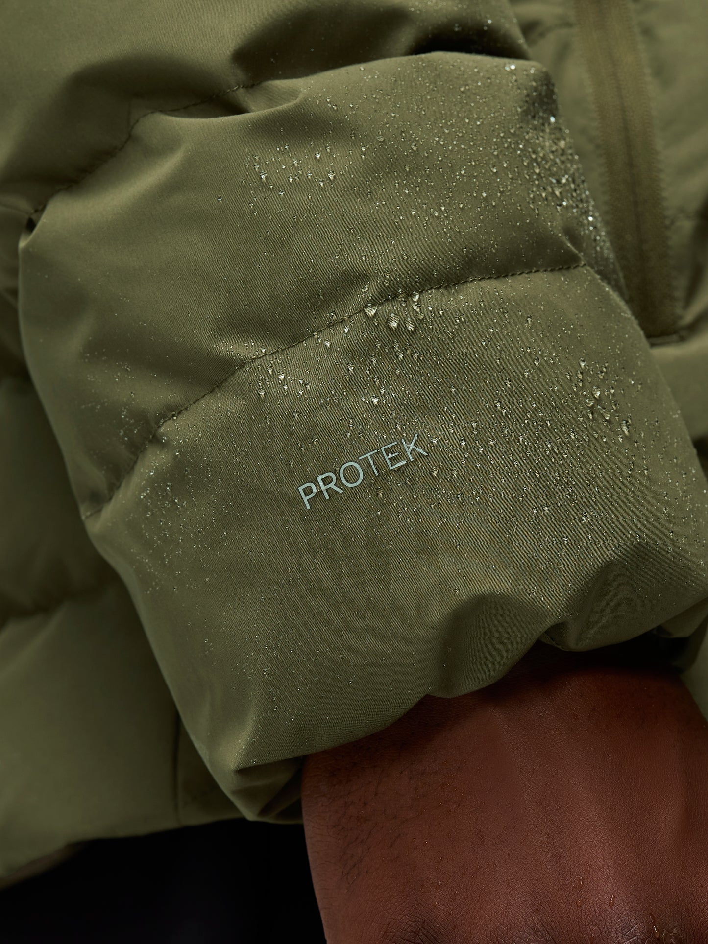 Close-up of olive green lightweight jacket fabric with water droplets and ProTek branding on the sleeve