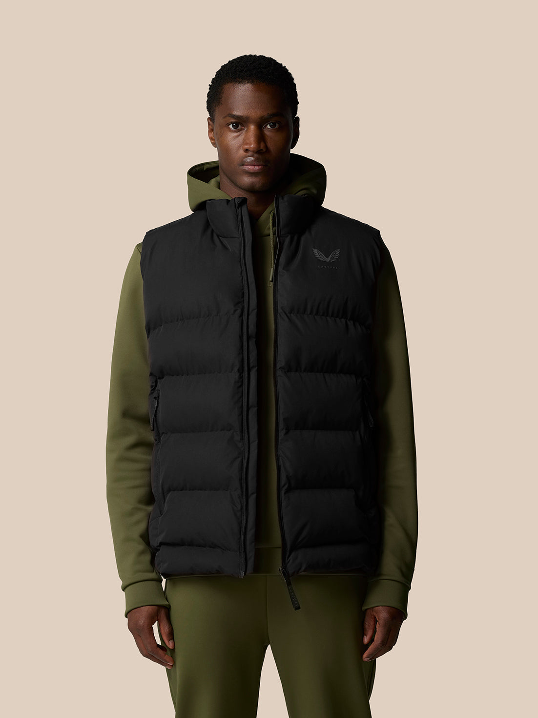 Male model wearing black sleeveless puffer gilet over olive green hoodie.