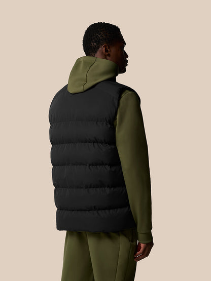 Back view of a man wearing a black sleeveless puffer gilet over an olive green hoodie.
