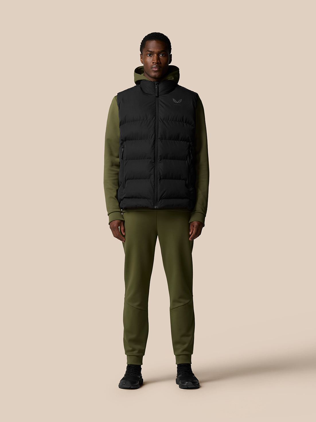 Male model wearing black sleeveless puffer gilet over an olive hoodie and olive joggers, front zip and zip pockets
