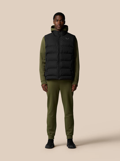 Male model wearing black sleeveless puffer gilet over an olive hoodie and olive joggers, front zip and zip pockets