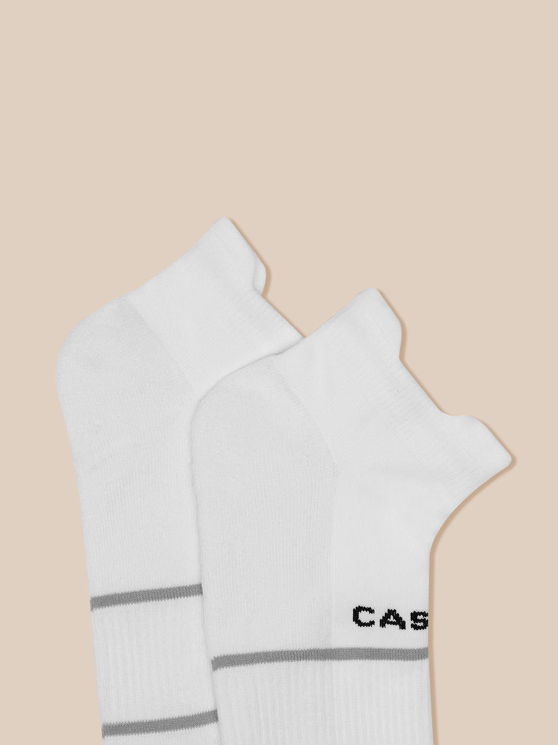 Two white ankle socks with gray bands and Castore branding on the cuffs against a beige backdrop