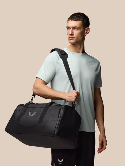 Man in a pale blue T shirt carries a black holdall over his shoulder with a crossbody strap