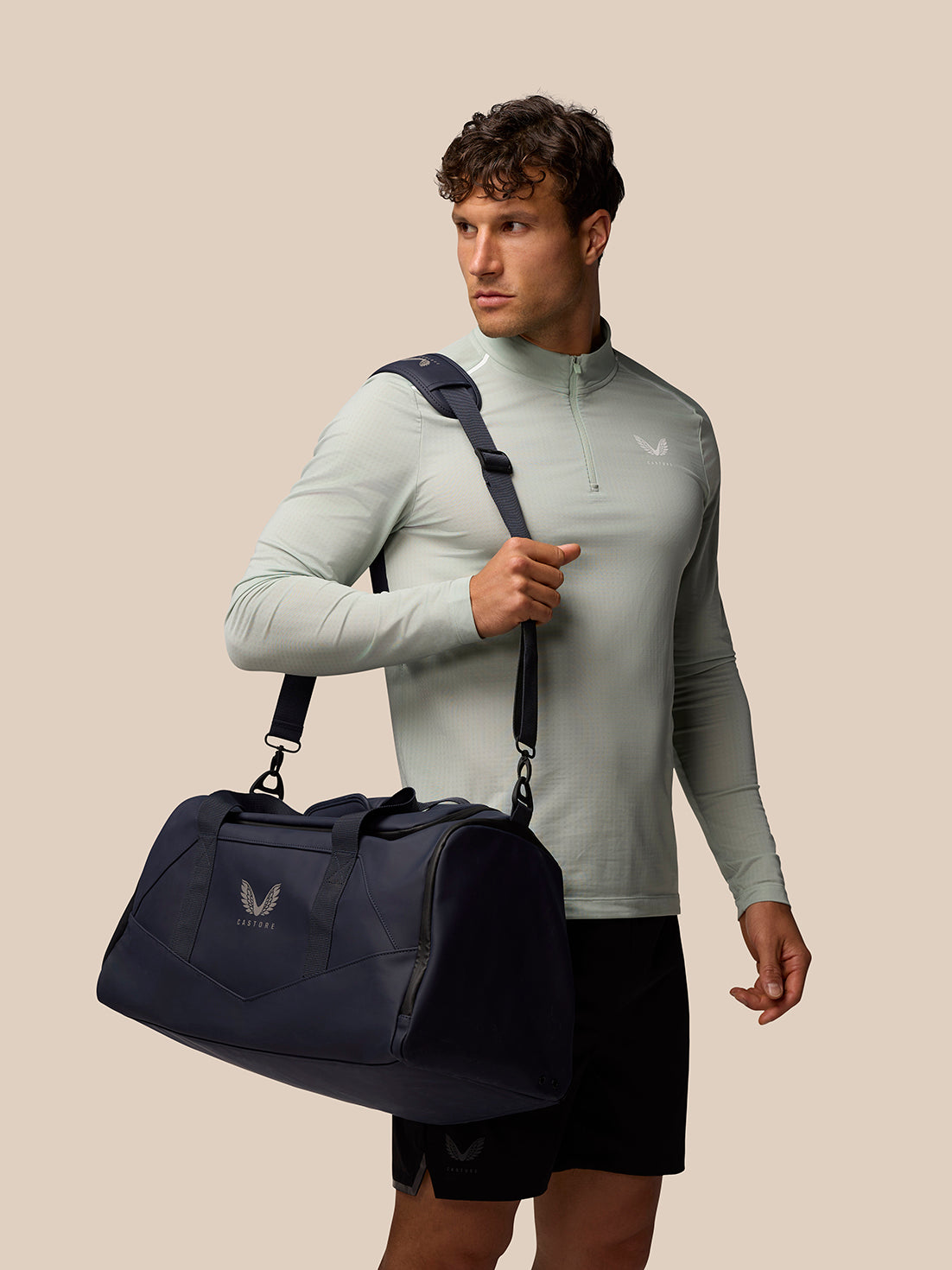 Male model carrying a navy holdall with logo and a detachable shoulder strap against a beige background.