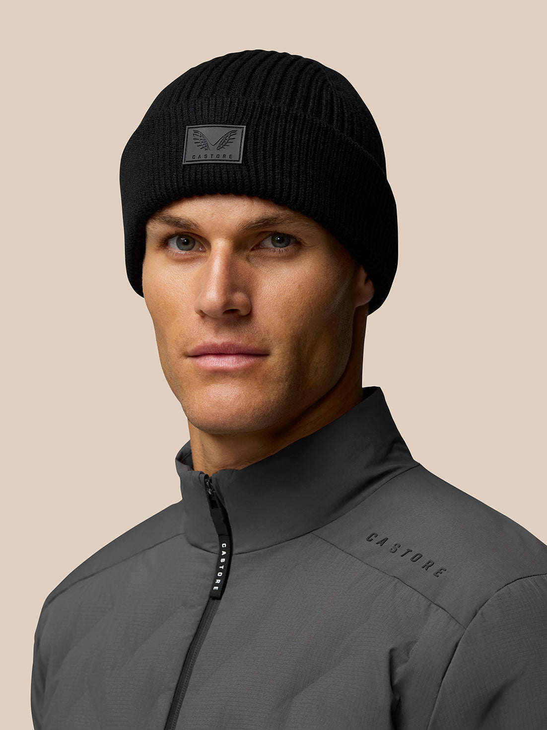 Model wearing black ribbed Castore beanie with front logo patch and grey jacket