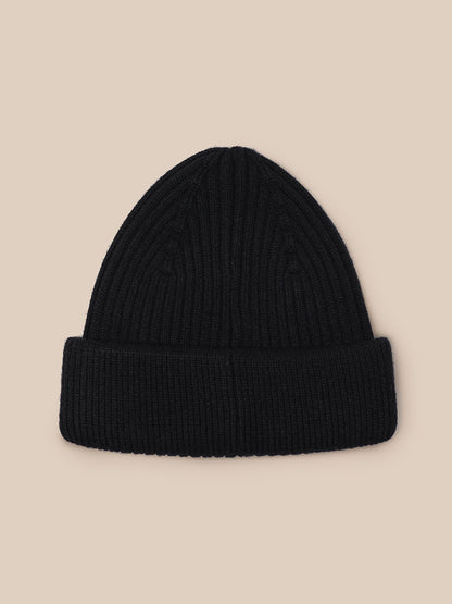 Close up of black ribbed knit beanie with folded cuff displayed on beige background.