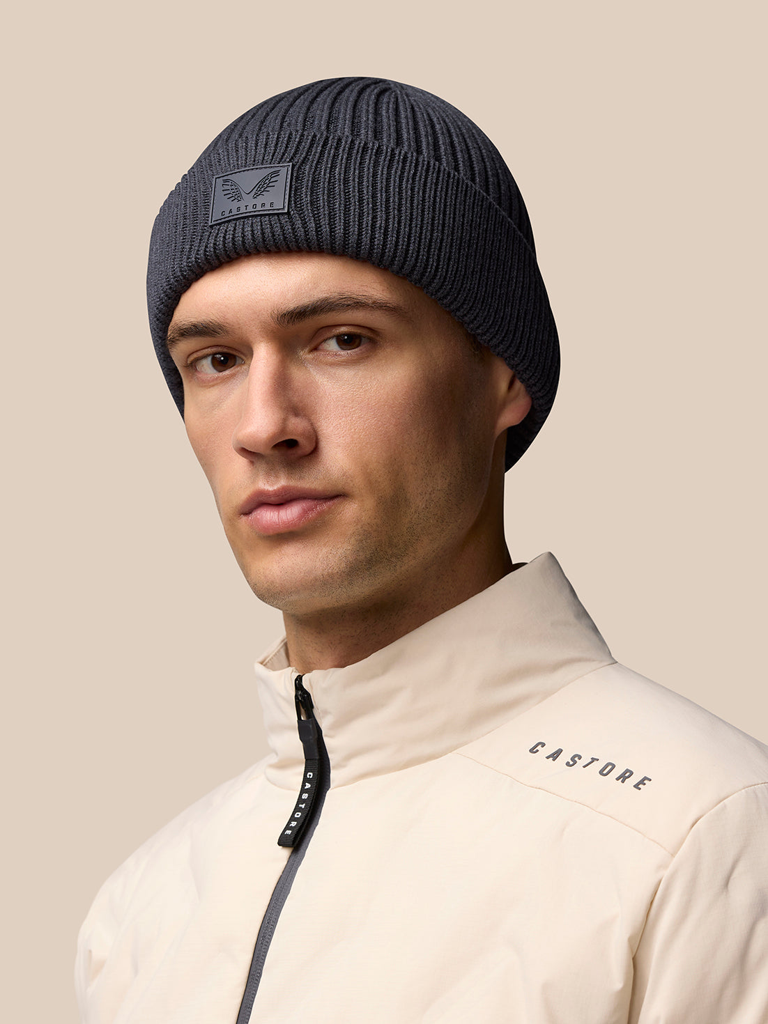 Grey ribbed Castore beanie with logo patch on front worn by male model in beige jacket.