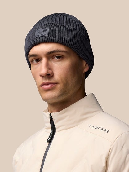 Grey ribbed Castore beanie with logo patch on front worn by male model in beige jacket.