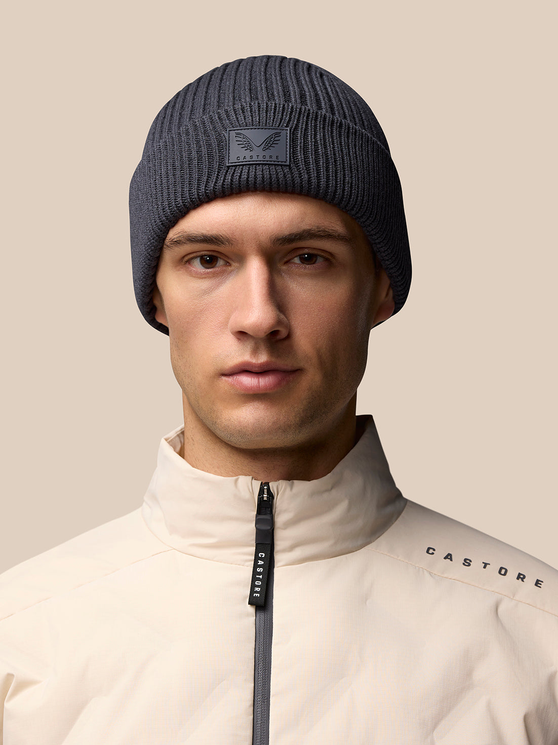 Dark grey ribbed Castore beanie with logo patch on front worn by male model in beige jacket.