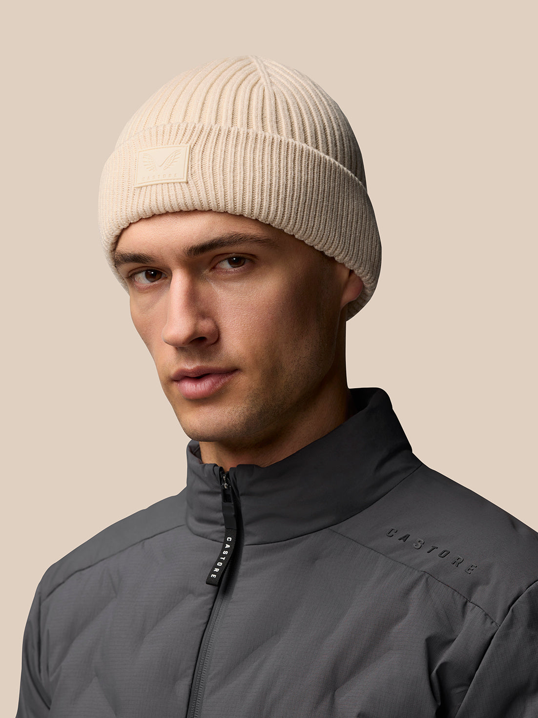 Beige Castore Active Beanie on a model, ribbed knit with front logo patch.