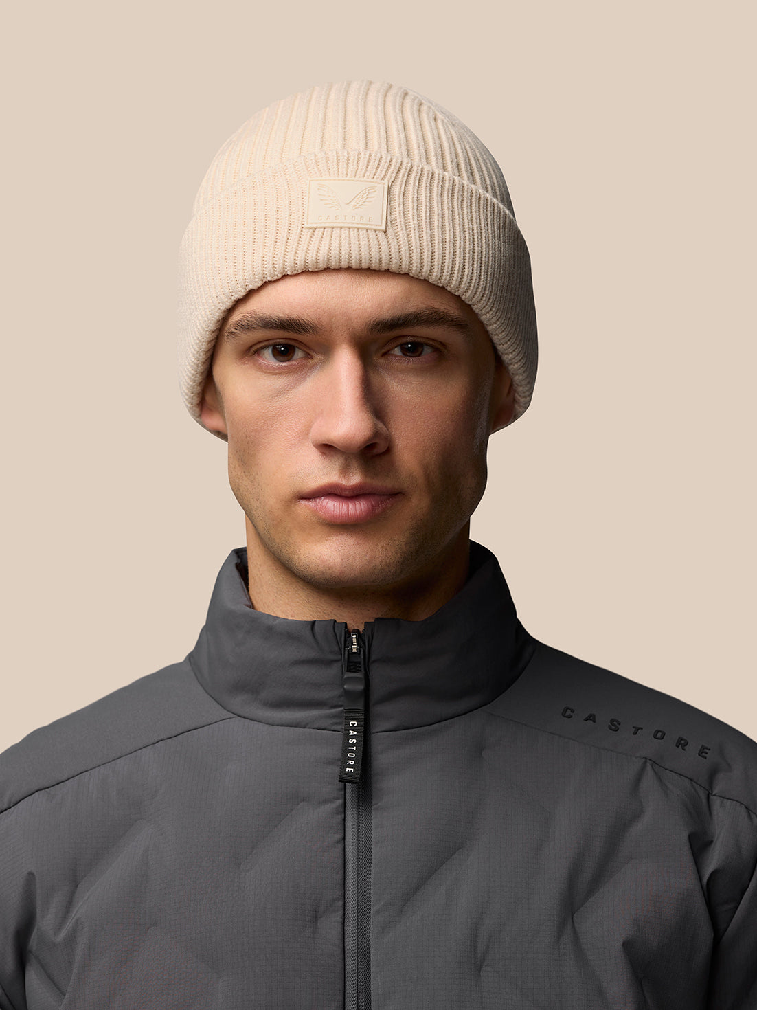 Beige Castore Active Beanie worn by a model in a gray jacket, ribbed knit with front logo.