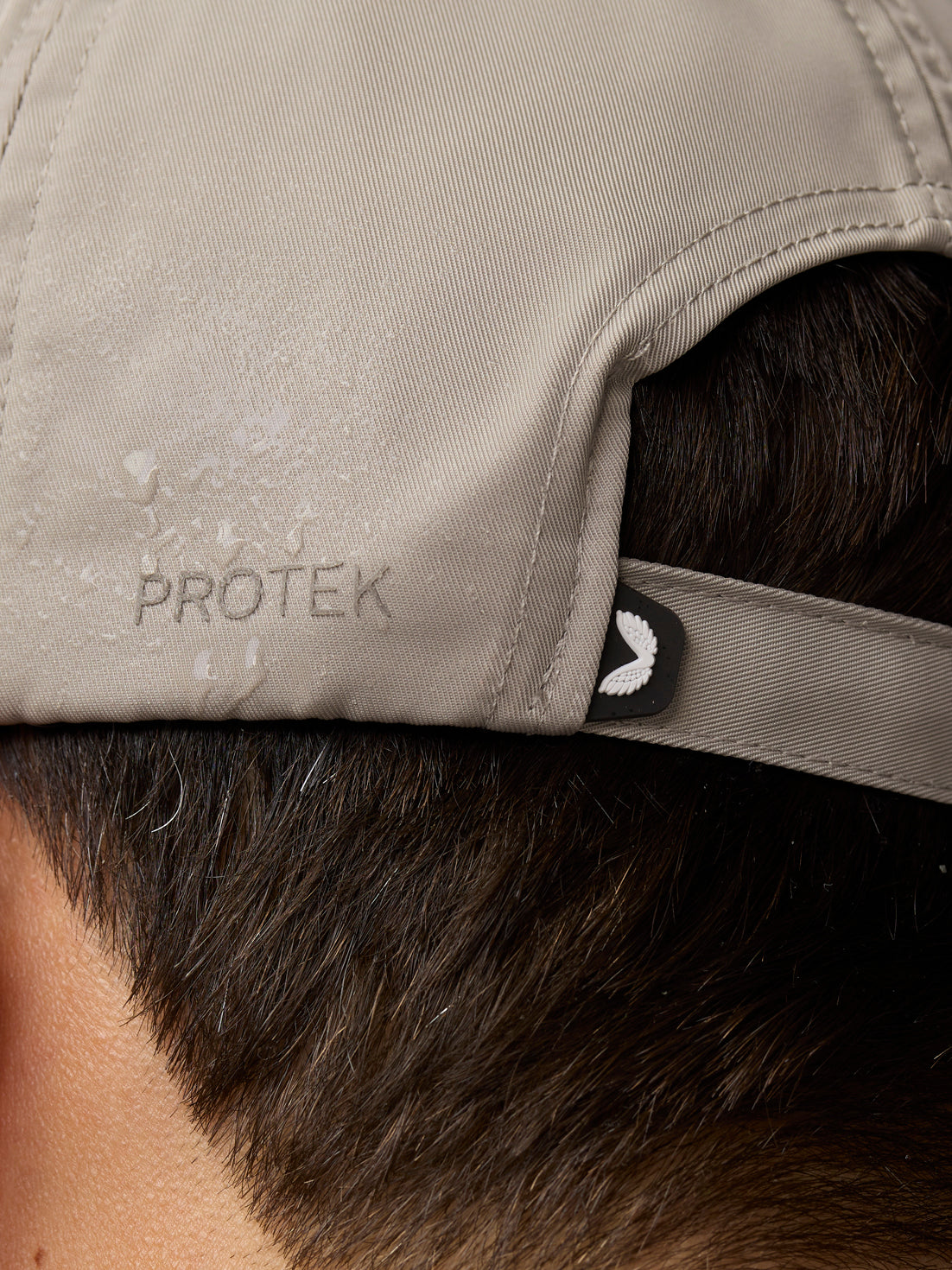 Close-up of rock grey cap fabric with PROTEK embossing and a small wing emblem at the back strap.