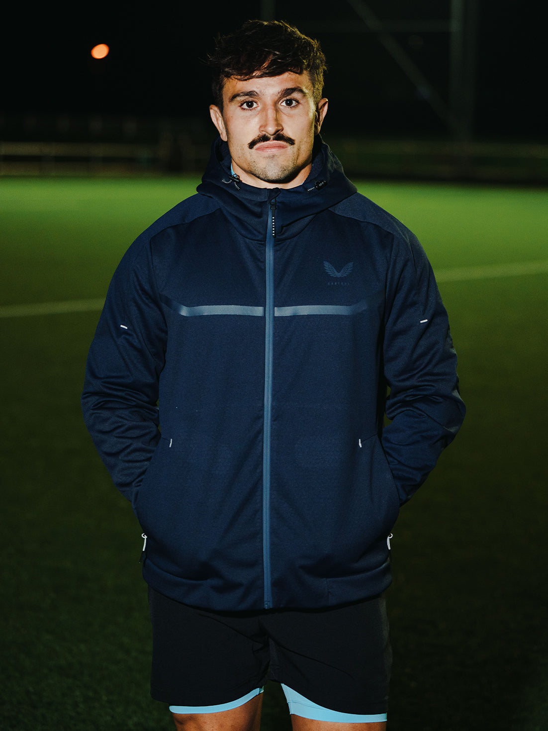 Male model wearing navy Garcia 3.0 Hoodie with hood on outdoors at night on a sports field