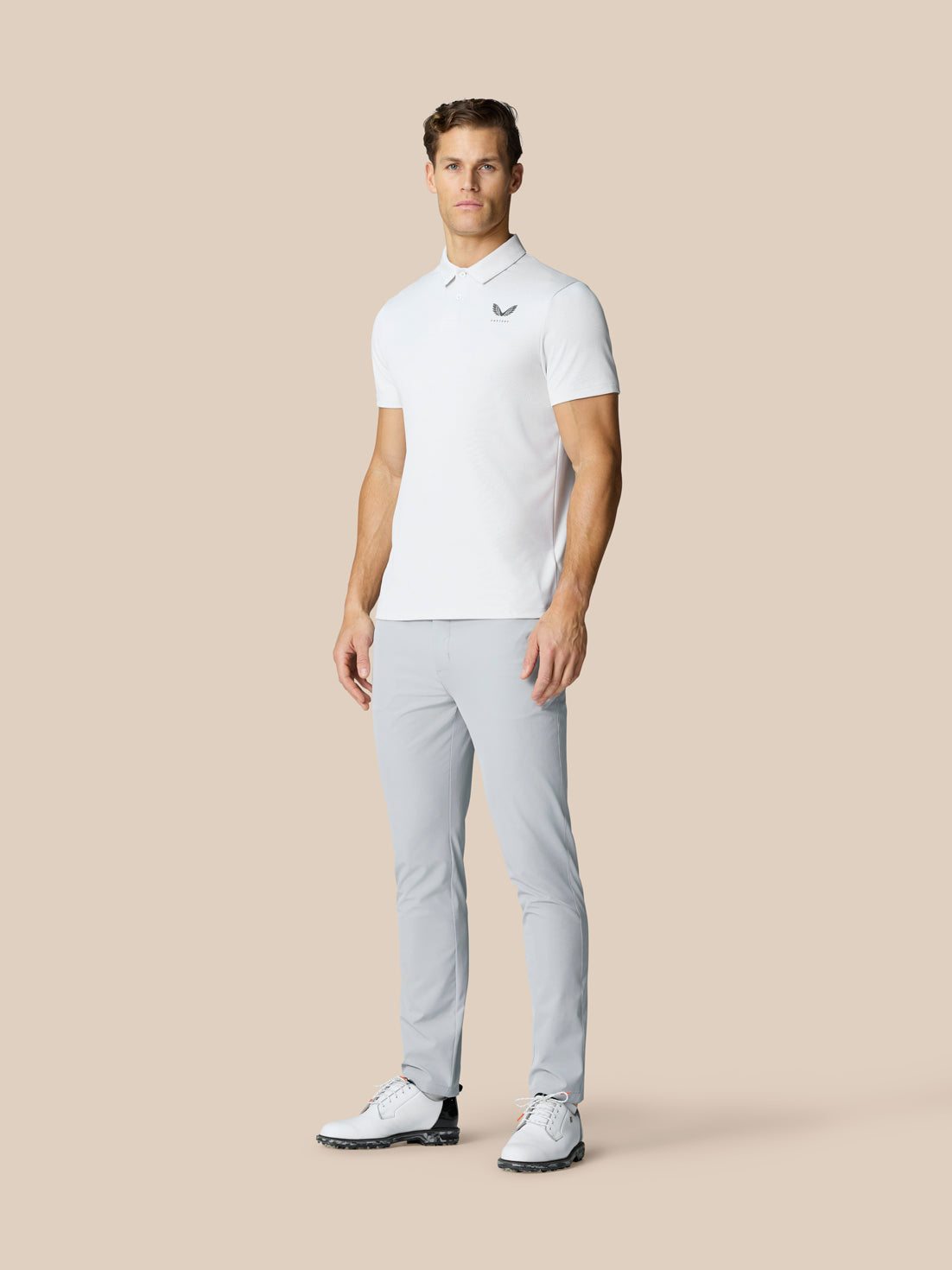 Full body shot of a man in light grey Castore Golf Club Trousers and a white polo, standing in golf shoes.
