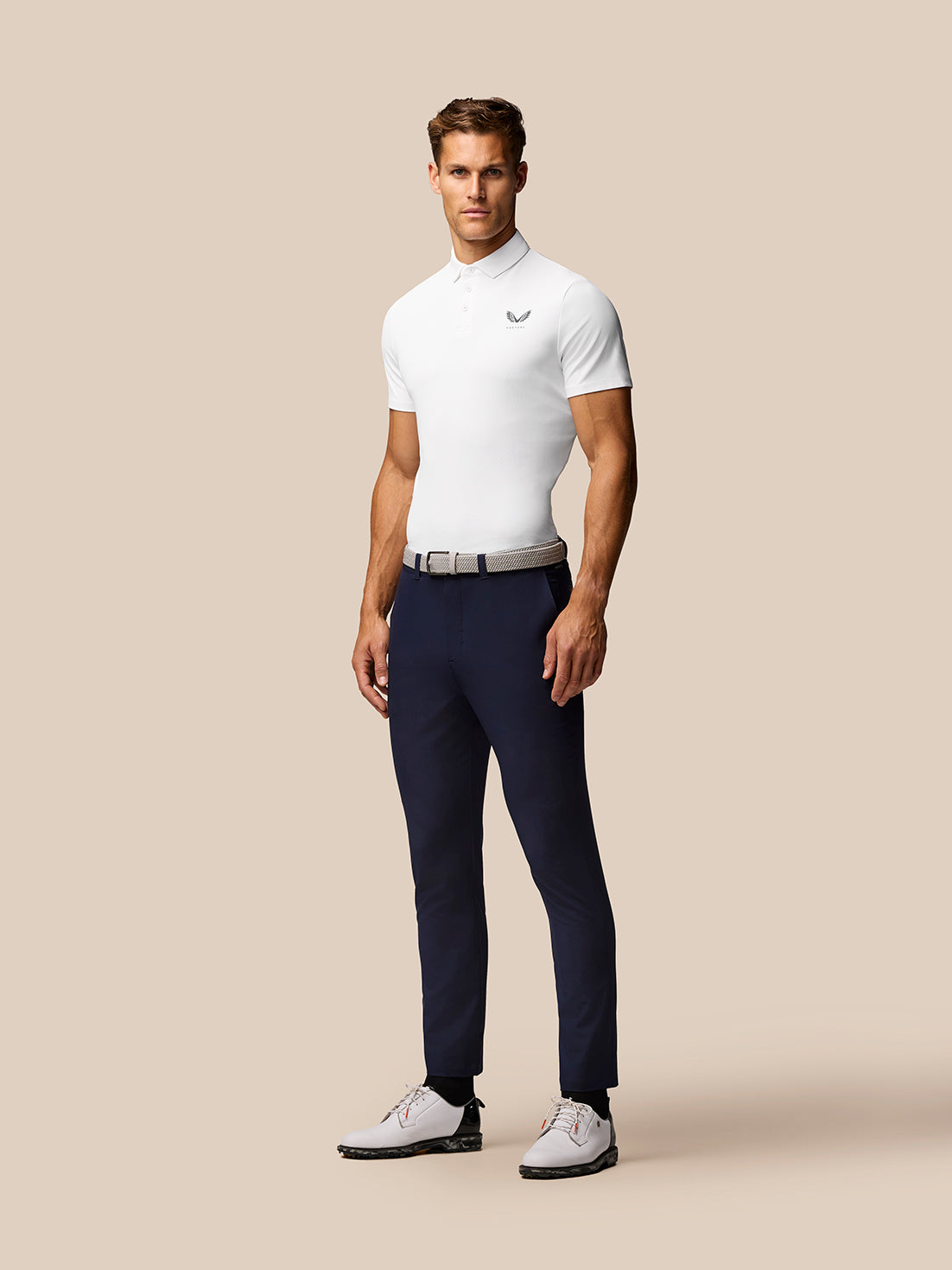 Male model wearing a white golf polo with Castore logo, navy trousers, belt, and white golf shoes against a beige background.